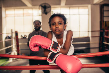 Confident looking boxing kid standing inside a boxing ring with her coach in the background. Girl wearing boxing gloves standing while resting her hands on the boxing ring. - JLPSF20341