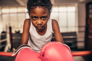 Kid wearing boxing gloves standing inside a boxing ring. Boxer kid resting her hands on boxing ring with determination and focus in her eyes. - JLPSF20342