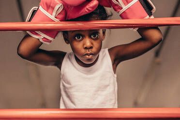 Kid wearing boxing gloves standing near a boxing ring. Close up of a kid boxer standing with hands above the head leaning on boxing ring. - JLPSF20345