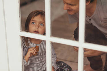 Kleines Mädchen sitzt am Fenster und schaut mit einem Stück Kuchen nach draußen. Mann sitzt neben seiner kleinen Tochter am Fenster. - JLPSF20351