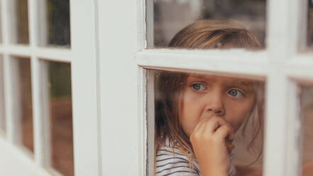 Nahaufnahme eines kleinen Mädchens, das am Fenster sitzt und nach draußen schaut. Ein Mädchen mit grauen Augen isst einen Kuchen und schaut aus dem Fenster. - JLPSF20352