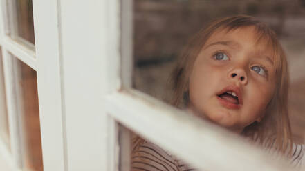 Close up of a little girl sitting beside a window and looking outside. Grey eyed girl looking outside sitting at the window. - JLPSF20353