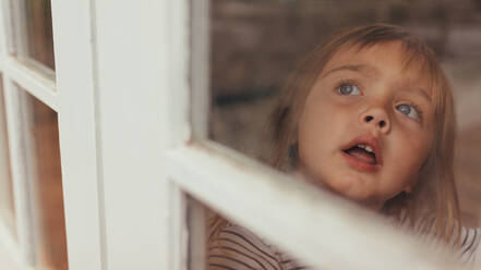 Nahaufnahme eines kleinen Mädchens, das am Fenster sitzt und nach draußen schaut. Grauäugiges Mädchen, das am Fenster sitzt und nach draußen schaut. - JLPSF20353