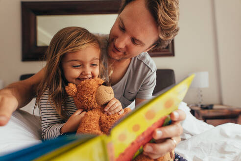 Vater und Tochter sitzen auf dem Bett und lesen eine Geschichte. Kleines Mädchen sitzt mit ihrem Vater, der einen Teddybär hält, und schaut sich ein Buch an. - JLPSF20359
