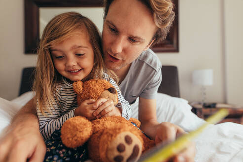 Vater und Tochter sitzen auf dem Bett und lesen eine Geschichte. Kleines Mädchen sitzt mit ihrem Vater, der einen Teddybär hält, und schaut sich ein Buch an. - JLPSF20360