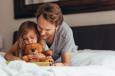Vater und Tochter sitzen auf dem Bett und lesen eine Geschichte. Kleines Mädchen sitzt mit ihrem Vater, der einen Teddybär hält, und schaut sich ein Buch an. - JLPSF20361