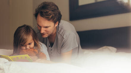 Father and daughter sitting on bed reading a story book. Little girl sitting with her father holding a teddy bear looking at a book. - JLPSF20362