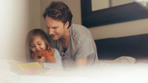 Vater und Tochter sitzen auf dem Bett und lesen eine Geschichte. Kleines Mädchen sitzt mit ihrem Vater, der einen Teddybär hält, und schaut sich ein Buch an. - JLPSF20363