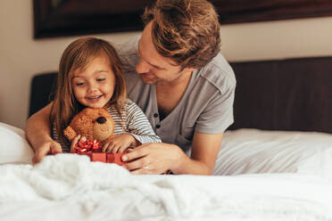 Vater und Tochter sitzen auf dem Bett und öffnen ein Geschenkpaket. Kleines Mädchen sitzt mit ihrem Vater, der einen Teddybär hält, und betrachtet ein Geschenkpaket. - JLPSF20364