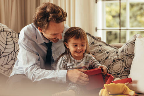 Vater und Tochter sitzen auf einem Sofa und öffnen ein Geschenkpaket. Der Vater hilft seiner Tochter beim Öffnen eines Geschenkpakets. - JLPSF20367