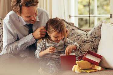 Vater und Tochter sitzen auf einem Sofa und öffnen ein Geschenkpaket. Das kleine Mädchen erkundet das Geschenkpaket, während ihr Vater zusieht. - JLPSF20368