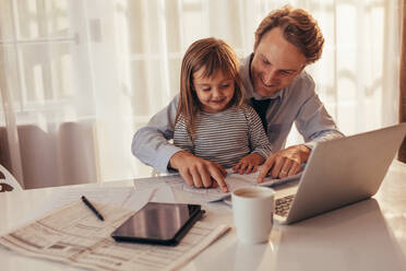 Mann sitzt mit seiner Tochter und schaut sich ein Buch mit Laptop, Tablet und Kaffeetasse auf dem Tisch an. Vater verbringt Zeit mit seiner Tochter, während er zu Hause arbeitet. - JLPSF20371