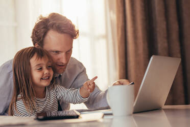 Ein Mann sitzt mit seiner Tochter vor einem Laptop mit einem Tablet und einer Kaffeetasse auf dem Tisch. Ein Vater verbringt Zeit mit seiner Tochter, während er zu Hause arbeitet. - JLPSF20373