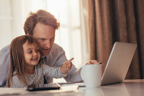 Ein Mann sitzt mit seiner Tochter vor einem Laptop mit einem Tablet und einer Kaffeetasse auf dem Tisch. Ein Vater verbringt Zeit mit seiner Tochter, während er zu Hause arbeitet. - JLPSF20373