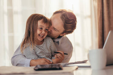 Man pampering his little daughter sitting with laptop computer and coffee cup on the table. Father spending time with daughter while working at home. - JLPSF20374