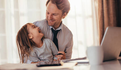 Man sitting with his daughter looking at her and smiling with laptop computer and coffee cup on the table. Father spending time with daughter while working at home. - JLPSF20375