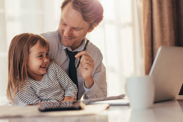 Man sitting with his daughter looking at her and smiling with laptop computer and coffee cup on the table. Father spending time with daughter while working at home. - JLPSF20376