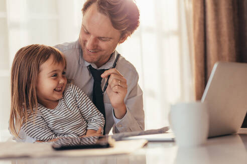 Mann sitzt bei seiner Tochter und schaut sie lächelnd an, mit Laptop und Kaffeetasse auf dem Tisch. Vater verbringt Zeit mit seiner Tochter, während er zu Hause arbeitet. - JLPSF20376