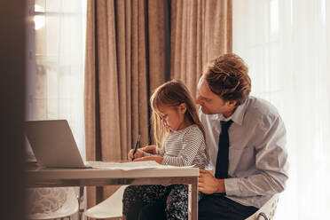 Kleines Mädchen schreibt mit Stift auf dem Schoß ihres Vaters mit Laptop auf dem Tisch. Vater verbringt Zeit mit Tochter bei der Arbeit zu Hause. - JLPSF20379