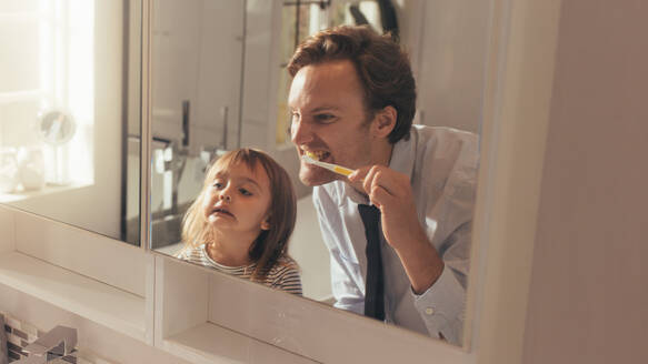 Man brushing his teeth looking into the mirror standing in bathroom while his daughter looks on. Man teaching his daughter how to brush teeth. - JLPSF20380
