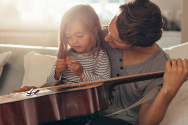 Mann sitzt mit seiner Tochter zu Hause und hält eine Gitarre. Mädchen sitzt auf dem Schoß ihres Vaters und hält ein Gitarrenplektrum. - JLPSF20384