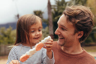 Glückliche Tochter mit ihrem Vater, der eine Zuckerstange im Freien hält. Vater und Tochter verbringen Zeit miteinander und essen Süßigkeiten. - JLPSF20395