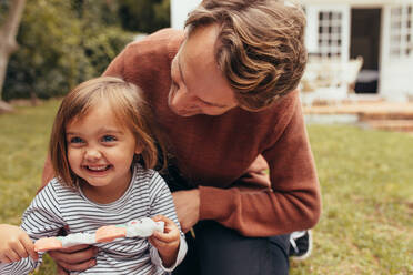 Lächelndes Mädchen sitzt mit ihrem Vater im Freien und hält eine Zuckerstange in der Hand. Vater und Tochter verbringen Zeit miteinander und essen Süßigkeiten. - JLPSF20396