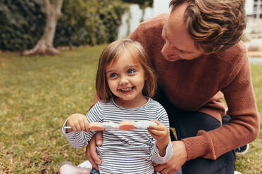 Lächelndes Mädchen sitzt mit ihrem Vater im Freien und hält eine Zuckerstange in der Hand. Vater und Tochter verbringen Zeit miteinander und essen Süßigkeiten. - JLPSF20397