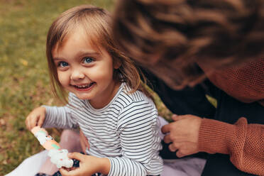 Lächelndes Mädchen sitzt mit ihrem Vater im Freien und hält eine Zuckerstange in der Hand. Vater und Tochter verbringen Zeit miteinander und essen Süßigkeiten. - JLPSF20398