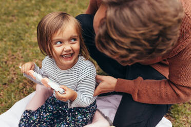 Lächelndes Mädchen sitzt mit ihrem Vater im Freien und hält eine Zuckerstange in der Hand. Vater und Tochter verbringen Zeit miteinander und essen Süßigkeiten. - JLPSF20399