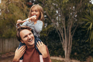 Smiling girl sitting on the shoulders of her father eating a sugar candy stick. Father and daughter spending time together eating sugar candy. - JLPSF20401