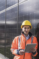 Smiling female engineer with tablet PC leaning on solar panels - UUF27689