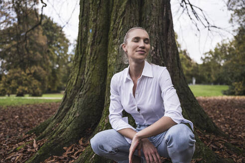 Lächelnde Frau mit geschlossenen Augen, die vor einem Baum hockt - UUF27704