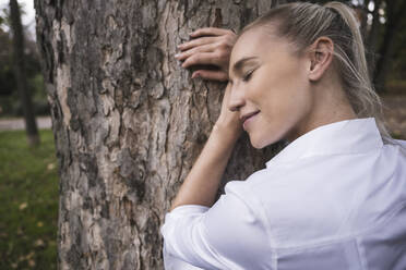 Smiling young woman with eyes closed leaning on tree - UUF27706
