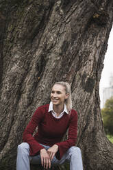 Glückliche junge Frau sitzt vor einem Baum - UUF27710