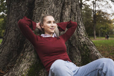 Smiling woman with hands behind head leaning on tree - UUF27712