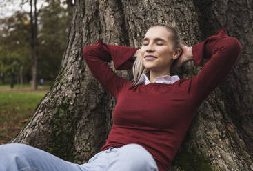 Smiling woman with hands behind head and eyes closed leaning on tree - UUF27713