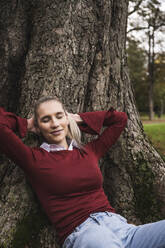Smiling young woman relaxing by tree at park - UUF27714