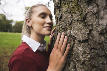 Smiling young woman standing with eyes closed by tree - UUF27717