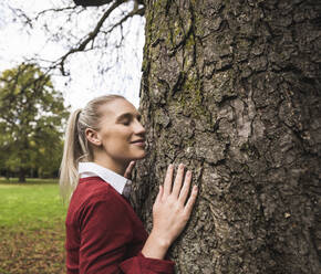 Smiling woman standing with eyes closed by tree at park - UUF27718