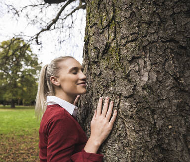Lächelnde Frau steht mit geschlossenen Augen an einem Baum im Park - UUF27718