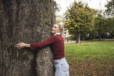 Smiling woman hugging tree at park - UUF27722