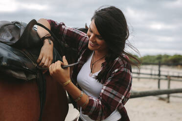 Eine Frau befestigt den Sattel eines braunen Pferdes. Eine Reiterin schnallt den Gurt auf den Sattel, während sie das Pferd im Stall vorbereitet. - JLPSF20403