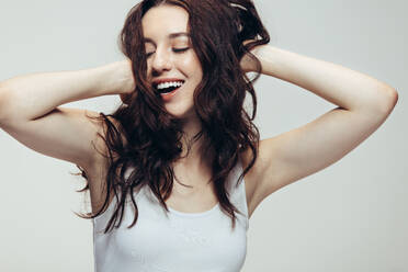 Woman in happy mood laughing with eyes closed and hands in hair. Close up of a young woman in casuals on grey background. - JLPSF20413