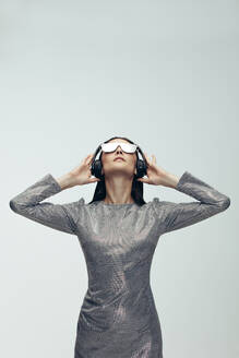Vertikale Aufnahme einer kaukasischen Frau, die Kopfhörer trägt und über einen grauen Hintergrund nach oben schaut. Stylisches weibliches Modell im Roboter-Look. - JLPSF20425