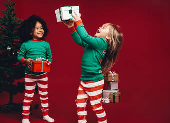 Kleine Mädchen haben Spaß beim Spielen mit den Geschenkboxen. Glückliches Kind hebt ihre Geschenkbox vor Freude, während ein anderes Kind mit einem Weihnachtsbaum im Hintergrund zusieht. - JLPSF20432