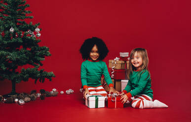 Kleine Mädchen spielen mit ihren Weihnachtsgeschenken auf dem Boden sitzend. Glückliche multiethnische Kinder mit ihren Weihnachtsgeschenken sitzen neben einem Weihnachtsbaum. - JLPSF20434