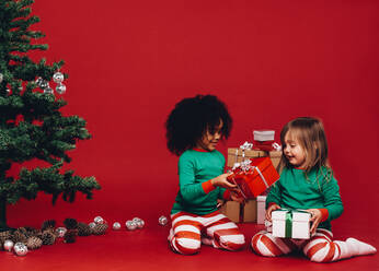 Multiethnische Kinder betrachten ihre Weihnachtsgeschenkboxen. Glückliche kleine Kinder mit ihren Weihnachtsgeschenken sitzen neben einem Weihnachtsbaum. - JLPSF20435