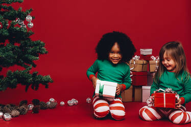 Glückliche Kinder spielen mit ihren Weihnachtsgeschenken auf dem Boden sitzend. Multi ethnische Kinder halten ihre Weihnachtsgeschenke sitzen neben einem Weihnachtsbaum. - JLPSF20436