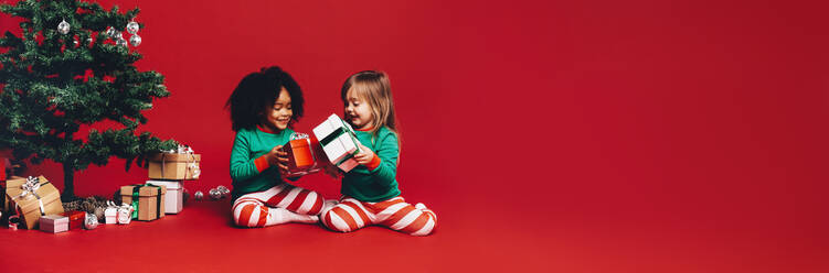 Kleine Mädchen sitzen neben einem Weihnachtsbaum und spielen mit ihren Geschenkboxen. Kinder tauschen Weihnachtsgeschenke auf rotem Hintergrund aus. - JLPSF20438